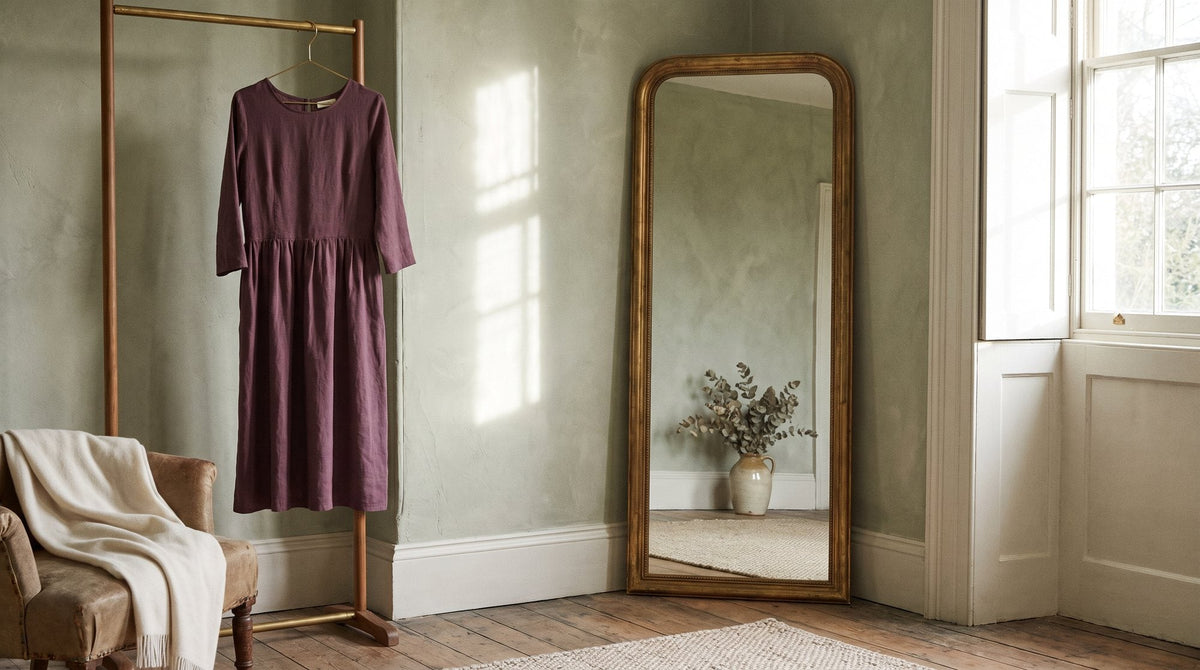 A full-length antique mirror leaning against a sage-green wall with a draped midi dress on a hanger beside it, no reflection of any person Image