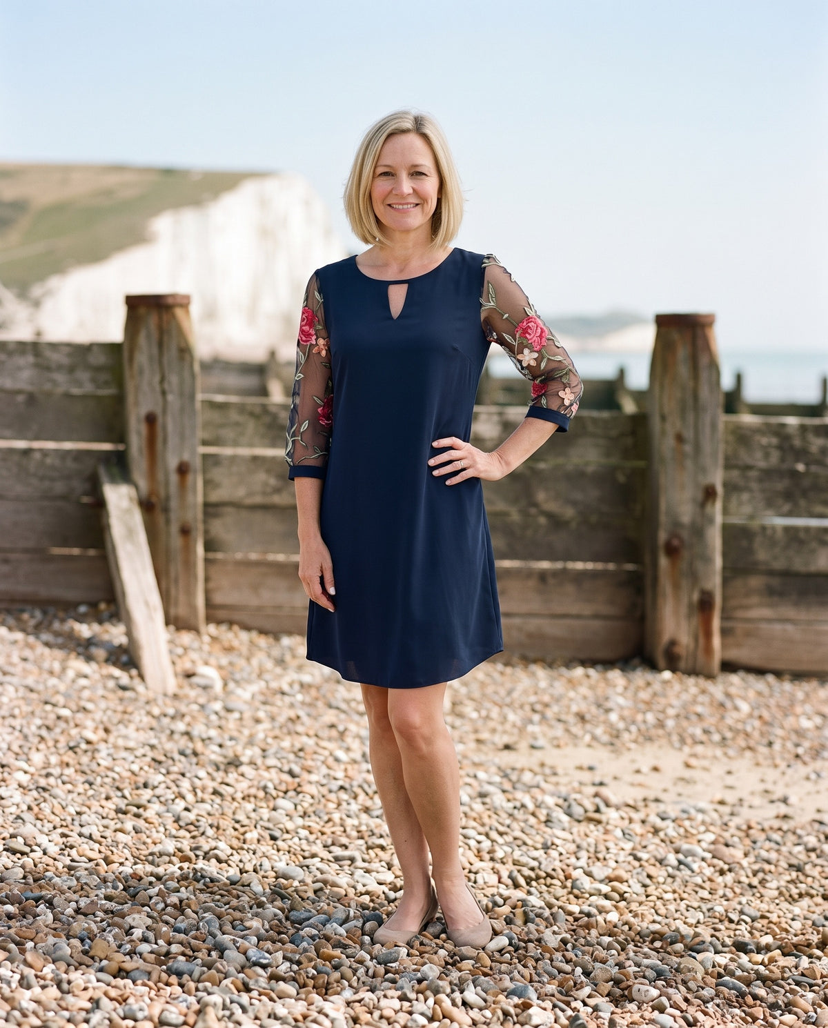 Woman wearing the shift dress with embroidered sheer sleeves on an English pebble beach